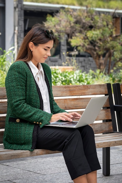 Mujer sonriente joven que trabaja con su computadora portátil desde la calle sentada en un banco de madera