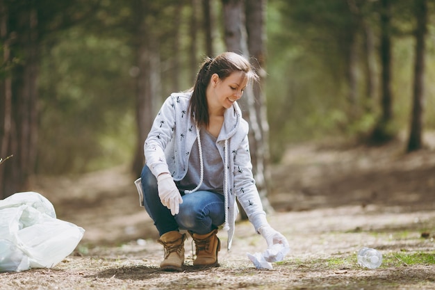 Mujer sonriente joven en ropa casual, guantes de limpieza de basura en bolsas de basura en el parque. Problema de la contaminación ambiental