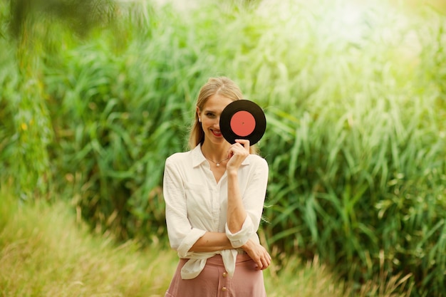 Mujer sonriente joven en ropa de estilo retro vintage tiene un disco de vinilo que cubre la mitad de la cara en el fondo de cañas en el lago