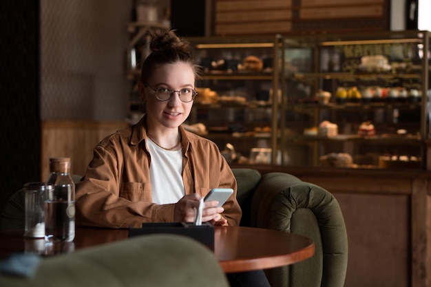 Mujer sonriente joven sentada en un café bebiendo café y usando un teléfono inteligente