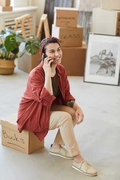 Mujer sonriente joven sentada en la caja con cosas y hablando por teléfono móvil durante la reubicación