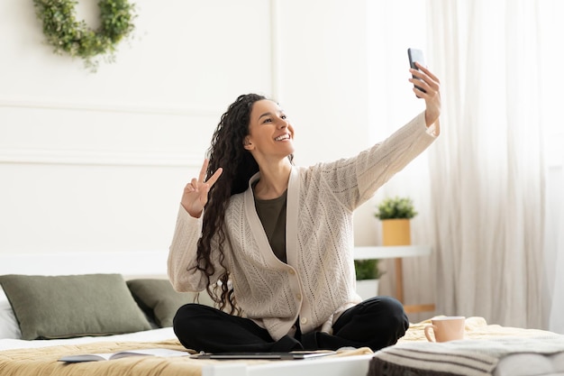 Mujer sonriente joven sentada en la cama tomando selfie