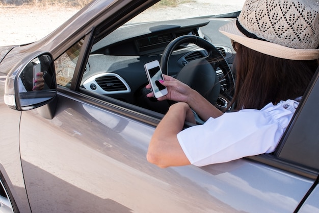 Mujer sonriente joven sentada en el coche inmóvil y escribiendo un mensaje mediante teléfono móvil