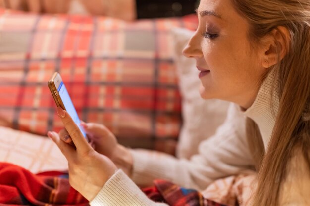 Mujer sonriente joven sincera se comunica con amigos usando un mensaje de texto de teléfono inteligente en casa en una cama acogedora en una manta a cuadros rojos o cuadros en feliz año nuevo y feliz nochebuena