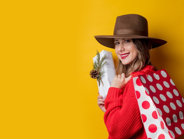 Mujer sonriente joven en suéter rojo con caja de regalo y bolsa de compras en la pared amarilla