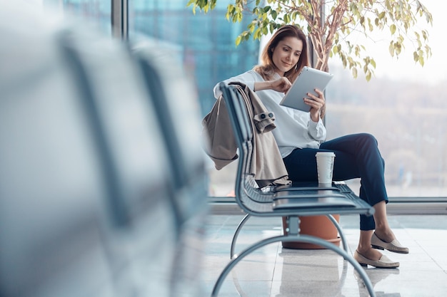 Mujer sonriente joven usando una tableta y esperando un vuelo en la sala de salidas del aeropuerto