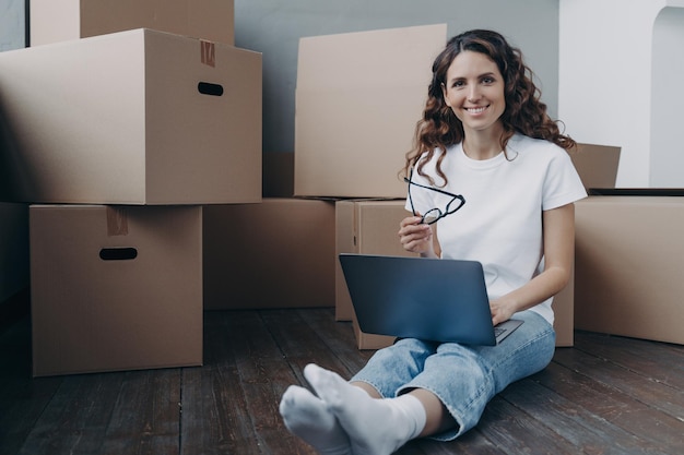 Mujer sonriente con laptop sentada en el piso cerca de cajas de cartón el día de la mudanza a la nueva casa