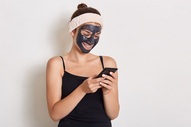 Mujer sonriente leyendo un mensaje en un teléfono inteligente con máscara negra en el rostro, vistiendo una banda para el cabello y una camiseta sin mangas