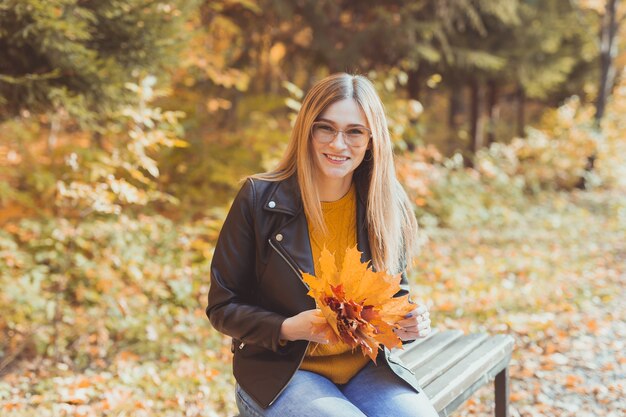 Mujer sonriente linda que sostiene las hojas de otoño en el parque de otoño. Concepto de temporada, estilo de vida y ocio.