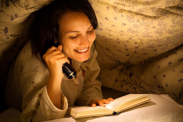 Foto mujer sonriente con linterna leyendo un libro en la cama bajo una manta