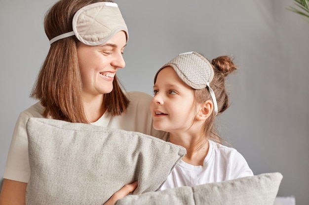 Mujer sonriente con máscara para dormir sentada en casa con su linda hijita en la cama hablando mirándose expresando emociones positivas