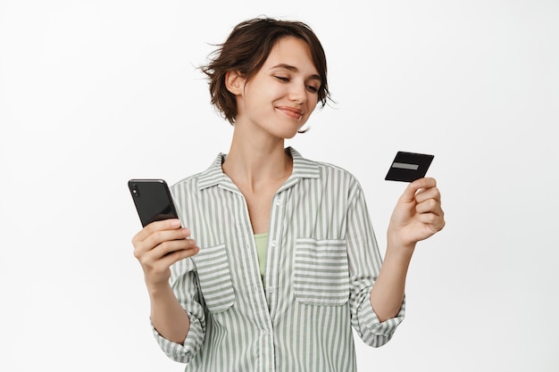 Foto mujer sonriente mirando satisfecho con tarjeta de crédito, sosteniendo el teléfono móvil, pagando en línea, haciendo compras, de pie en blanco