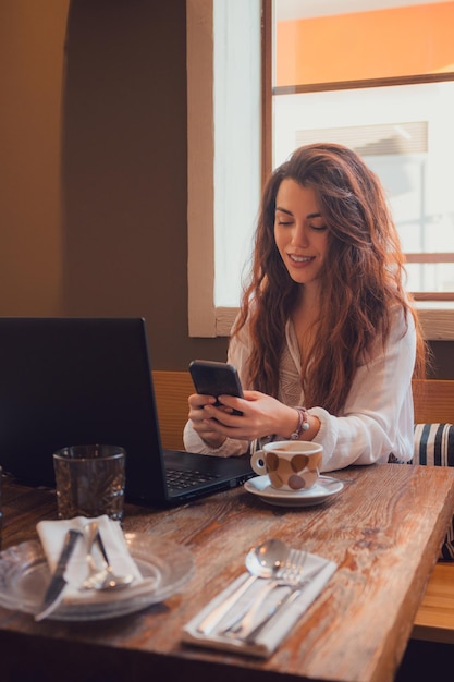 Mujer sonriente mirando su teléfono mientras trabaja desde un restaurante con una computadora portátil