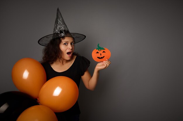 Mujer sonriente misteriosa sorprendida con sombrero de mago, vestida de negro, sostiene globos aerostáticos negros anaranjados y calabaza negra cortada a mano en fieltro, mira a la cámara, posa sobre fondo gris, espacio de copia.