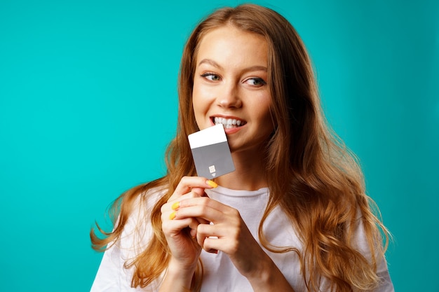 Mujer sonriente mordiendo una tarjeta de crédito en la tentación de ir de compras contra el fondo azul.