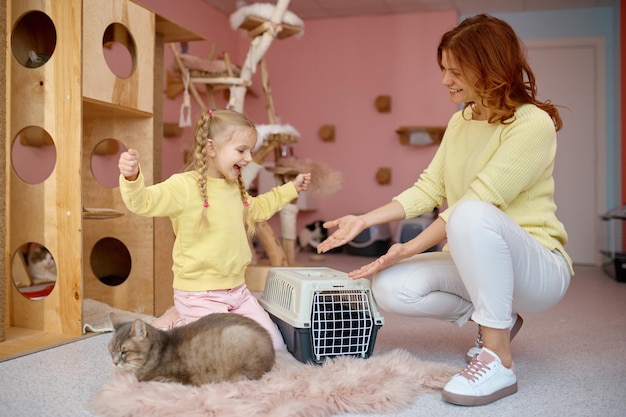 Mujer sonriente mostrando un gato en una jaula a una linda niña un poco asustada. Tiempo de diversión en un refugio para mascotas