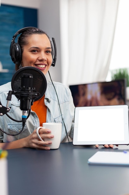 Mujer sonriente mostrando tableta digital con pantalla en blanco. Blogger que sostiene el dispositivo con plantilla de maqueta y fondo aislado mientras usa micrófono y auriculares para transmitir podcast.