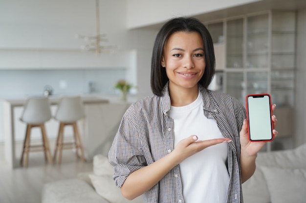 Mujer sonriente mostrar teléfono con pantalla de maqueta en blanco anunciar aplicaciones móviles en casa Oferta de tienda en línea