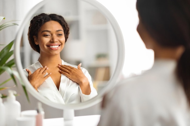 Mujer sonriente negra mirándose en el espejo y tocando la zona del escote