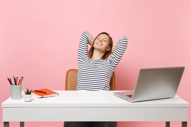 Mujer sonriente con los ojos cerrados descansando tomados de la mano detrás de la cabeza sentarse después del trabajo en el escritorio blanco con un ordenador portátil contemporáneo aislado sobre fondo rosa pastel. Logro de carrera empresarial. Copie el espacio.