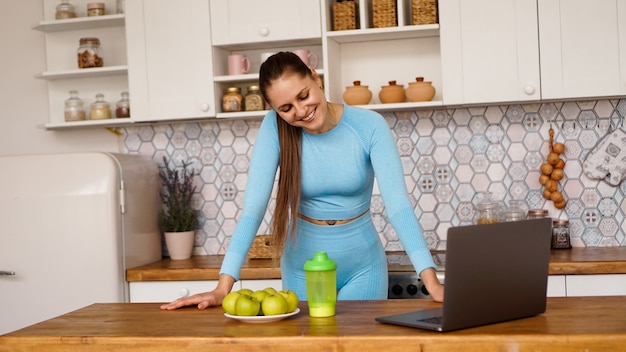 Mujer sonriente con ordenador en el interior de la cocina moderna. Concepto de estilo de vida saludable y cocina. Mujer transmite en línea y gesticula emocionalmente