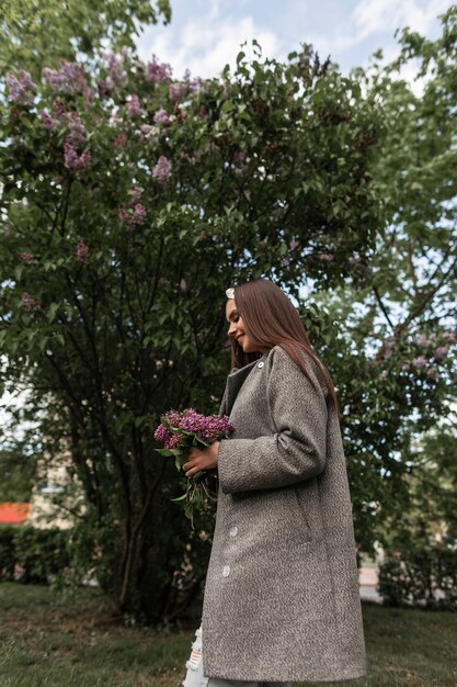 Mujer sonriente en pañuelo de moda en elegante abrigo gris tiene ramo de lilas en la mano cerca de árboles verdes. Feliz linda chica con dulce sonrisa en ropa de moda camina con hermosas flores en el parque.