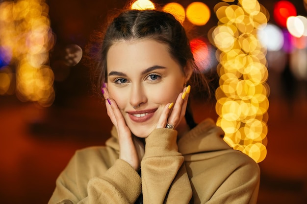 Mujer sonriente pararse fuera de la noche de invierno en la atmósfera de vacaciones guirnaldas de iluminación