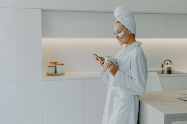 Mujer sonriente con parches de hidrogel debajo de los ojos disfruta de una bebida caliente mientras usa el teléfono móvil