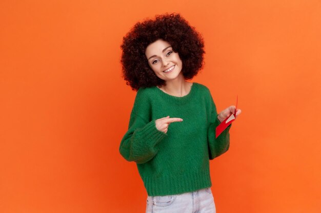 Mujer sonriente con peinado afro con suéter verde de estilo casual sosteniendo y señalando un sobre rojo en su mano carta romántica Estudio interior aislado en fondo naranja