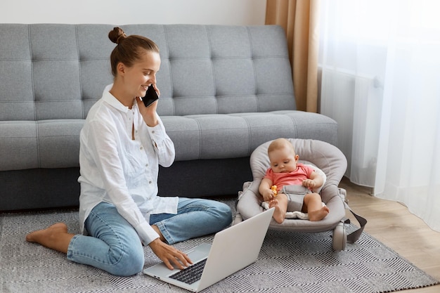 Mujer sonriente con peinado de moño con camisa blanca y jeans madre en licencia de maternidad haciendo trabajo en línea cuidando a su hija recién nacida y hablando por teléfono celular