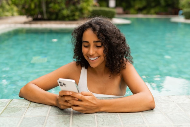 Mujer sonriente con peinado rizado haciendo autorretrato por teléfono móvil mientras se relaja en la piscina.