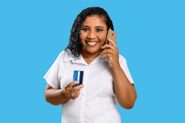 Una mujer sonriente de piel oscura sostiene una tarjeta de crédito y habla por teléfono celular aislada de fondo azul