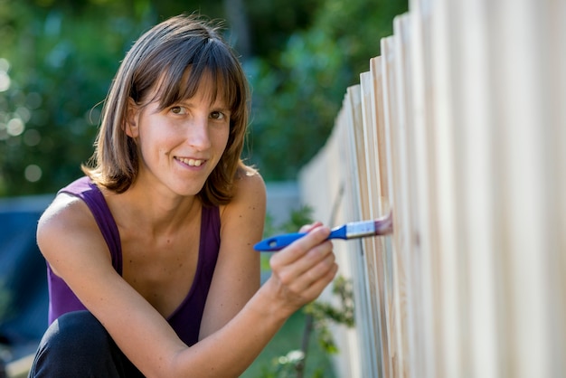 Mujer sonriente pintando una valla de jardín con un pincel en un concepto de bricolaje y mantenimiento de jardines.