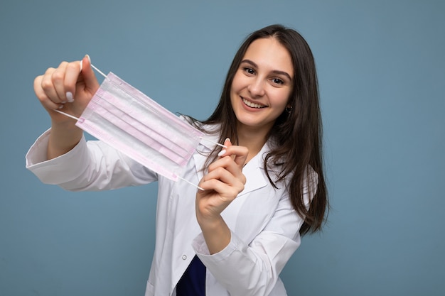 Mujer sonriente positiva con máscara protectora de virus contra el coronavirus y con bata médica blanca aislada sobre fondo azul.