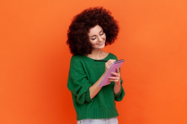 Mujer sonriente positiva con el pelo rizado y un suéter de estilo casual verde escribiendo en un cuaderno de papel haciendo una lista de tareas con buen humor Foto de estudio interior aislada en un fondo naranja