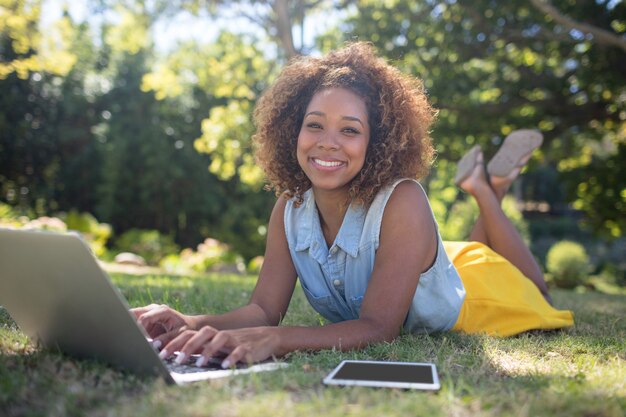 Mujer sonriente que miente en hierba y que usa la computadora portátil