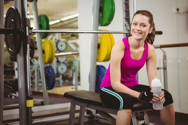 Mujer sonriente que se sienta en banco del barbell en el gimnasio