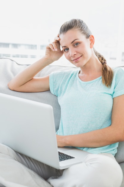 Mujer sonriente que se sienta en el sofá usando su computadora portátil