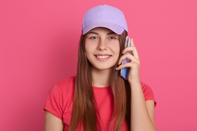 Mujer sonriente que sostiene el teléfono elegante cerca de su oído y que parece sonriente, vistiendo la camiseta casual roja y la gorra de béisbol, oponiéndose a la pared rosada.