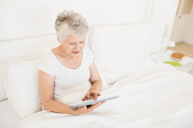 Mujer sonriente que usa la tableta que se sienta en la cama