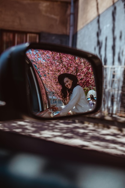 Mujer sonriente reflejo en el espejo retrovisor del coche floreciente árbol de sakura en el fondo