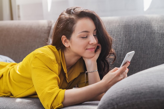 Mujer sonriente relajada en el sofá usando un teléfono inteligente y enviando mensajes de texto en casa