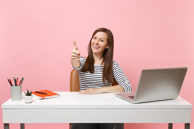 Mujer sonriente en ropa casual mostrar pulgar arriba sentarse trabajar en un escritorio blanco con portátil pc contemporáneo