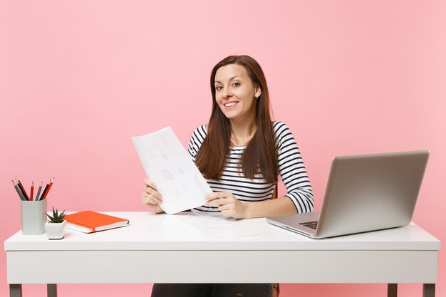 Mujer sonriente en ropa casual sosteniendo documentos en papel y trabajando en un proyecto mientras está sentado en la oficina con un portátil aislado sobre fondo rosa pastel. Concepto de carrera empresarial de logro. Copie el espacio.