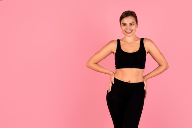 Mujer sonriente con ropa deportiva con las manos en las caderas posando sobre un fondo rosa