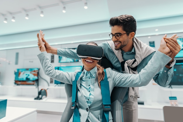 Mujer sonriente en ropa formal probando tecnología de realidad virtual mientras está sentado en la silla en la tienda de tecnología