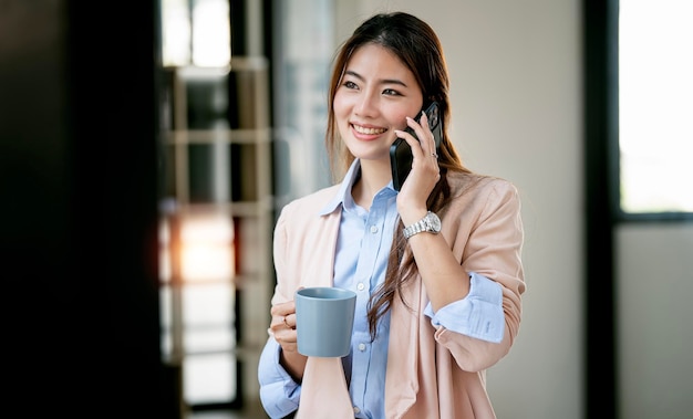 Mujer sonriente en ropa informal de pie en la oficina sosteniendo una taza y hablando por teléfono móvil