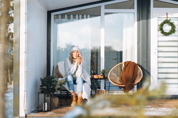 Mujer sonriente con ropa de invierno con taza de café sentada cerca de una casa de campo en invierno