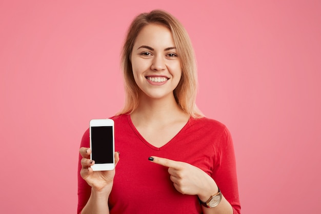 La mujer sonriente rubia alegre sostiene el teléfono celular moderno con la pantalla en blanco de la copia, indica en él, anuncia sus características, aislado en estudio rosado. Concepto de tecnología y personas