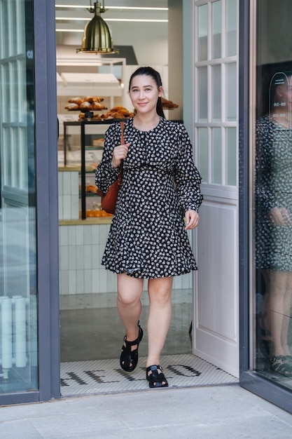 Mujer sonriente, saliendo del café. Lleva un vestido de verano negro con estampado de flores. Sosteniendo un abrigo en sus manos